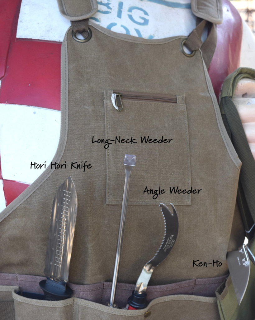 A brown work apron with labeled gardening tools in pockets: Hori Hori Knife, Long-Neck Weeder, Angle Weeder, and Ken-Ho. The apron is worn by a mannequin in front of a red and white object.