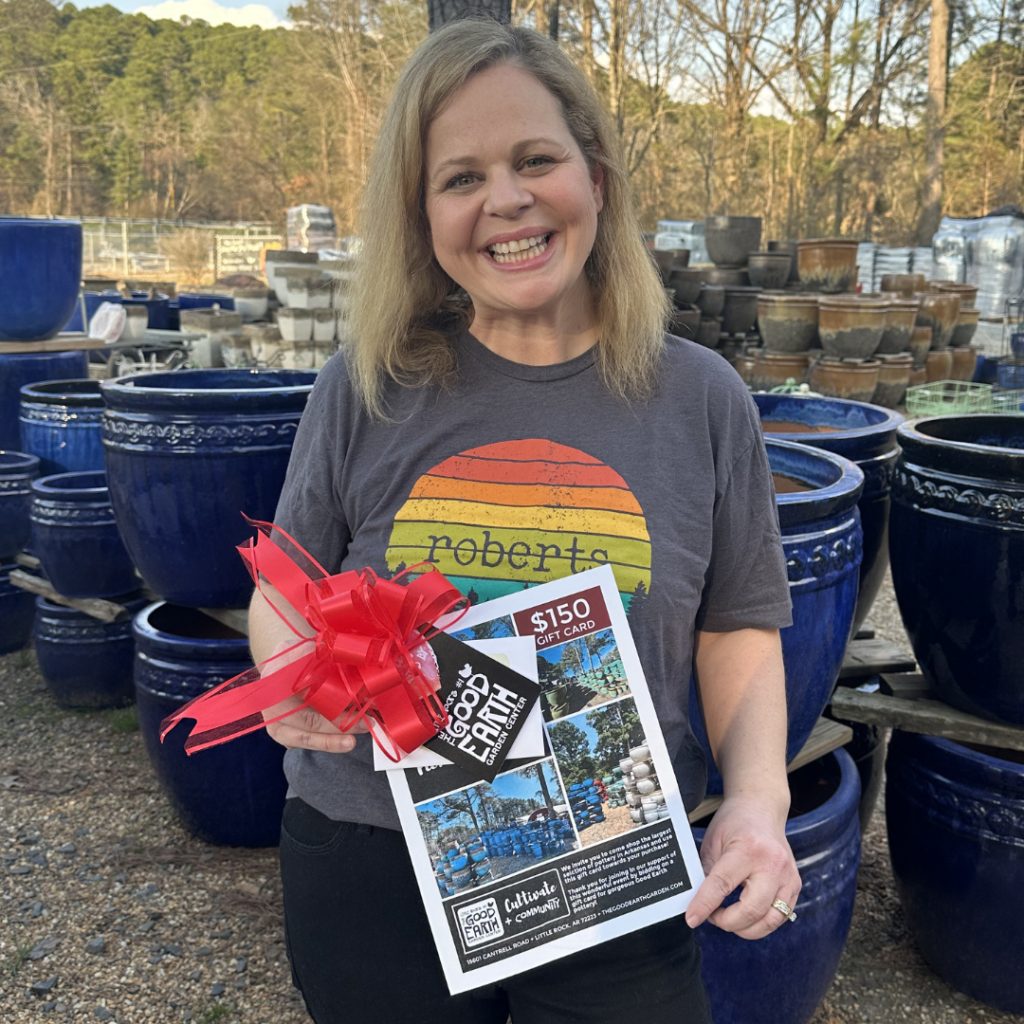 Smiling woman in a “Roberts” T-shirt holds a $150 gift card flyer and a red bow, standing outdoors among large blue ceramic pots with trees in the background.