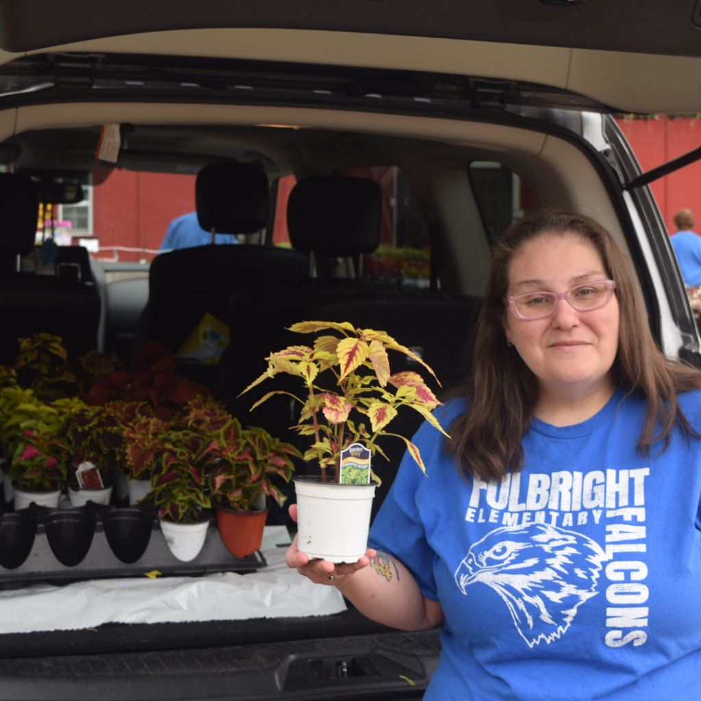 A woman wearing glasses and a Fulbright Falcons shirt holds a potted plant in front of an open car trunk filled with more plants.