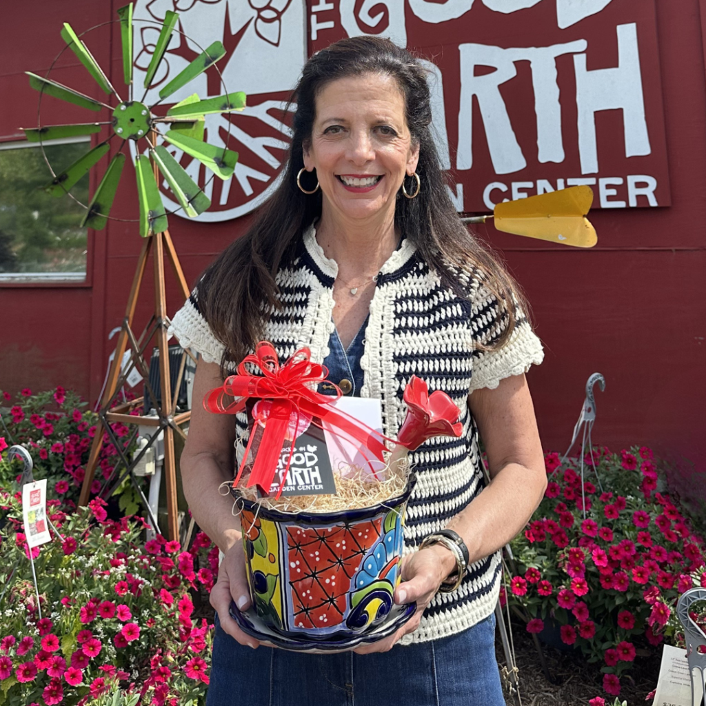 A woman in a black-and-white vest holds a colorful flower pot with a red bow, standing in front of a red wall with a Good Earth Garden Center sign and blooming pink flowers around her.