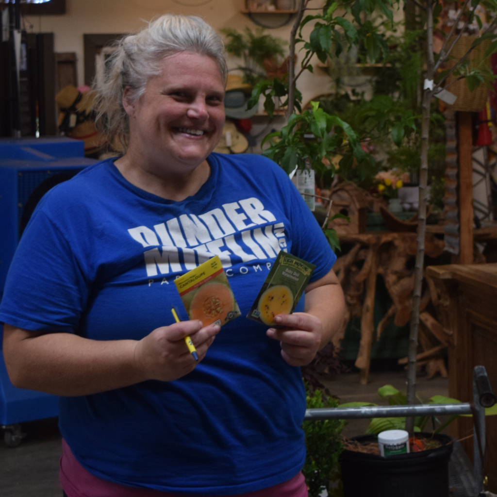 A smiling woman in a blue Bilder Museum shirt holds seed packets and a pencil while standing indoors near plants and gardening supplies.
