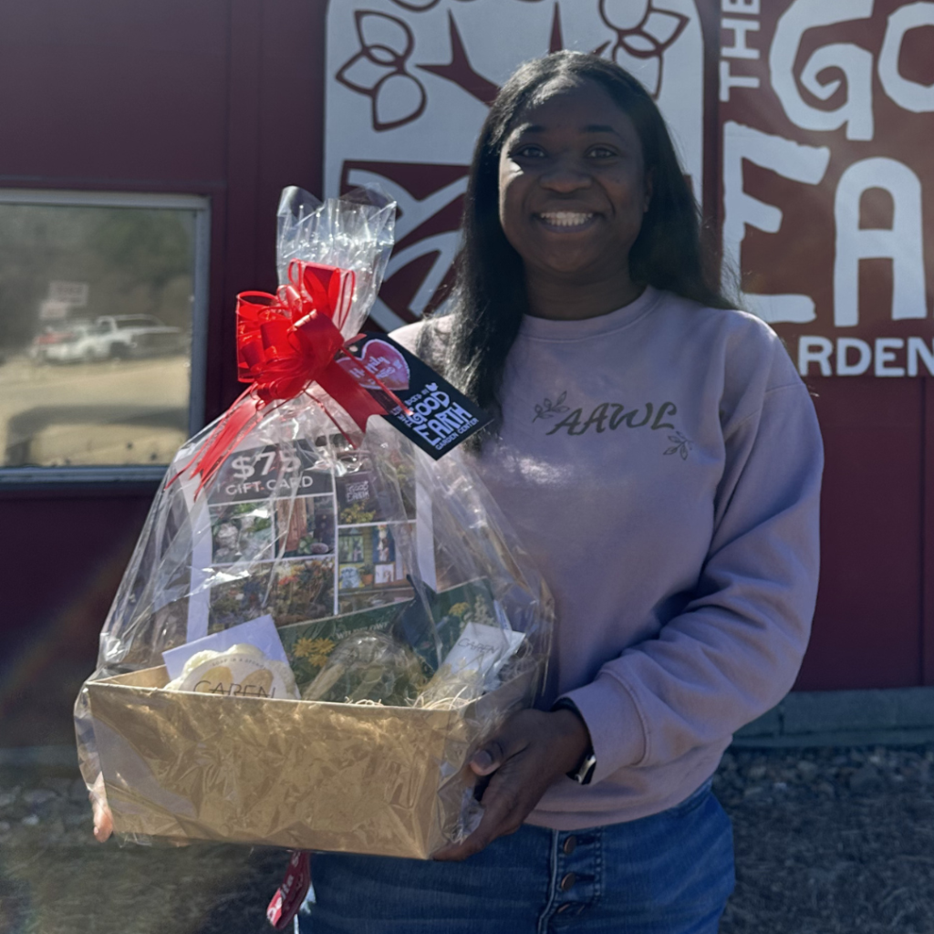 A smiling woman in a lavender sweatshirt holds a large gift basket wrapped in cellophane with a red bow. She stands outside in front of a building with signage that reads The Good Earth Garden.