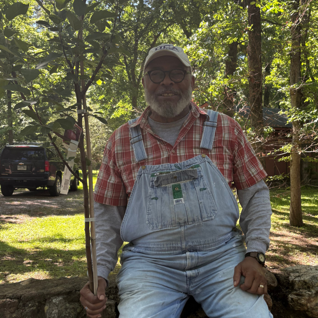 A smiling man in overalls and a plaid shirt sits on a stone wall outdoors, holding a young tree. Trees and a car are visible in the background on a sunny day.