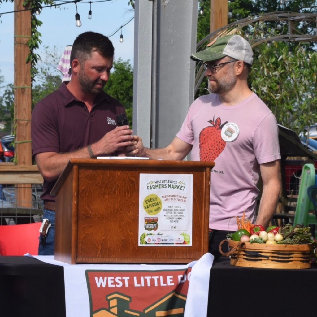 Two men stand at a podium at an outdoor farmers market event. One hands a microphone to the other. The podium has a flyer and produce is displayed nearby. A West Little Rock banner is visible.