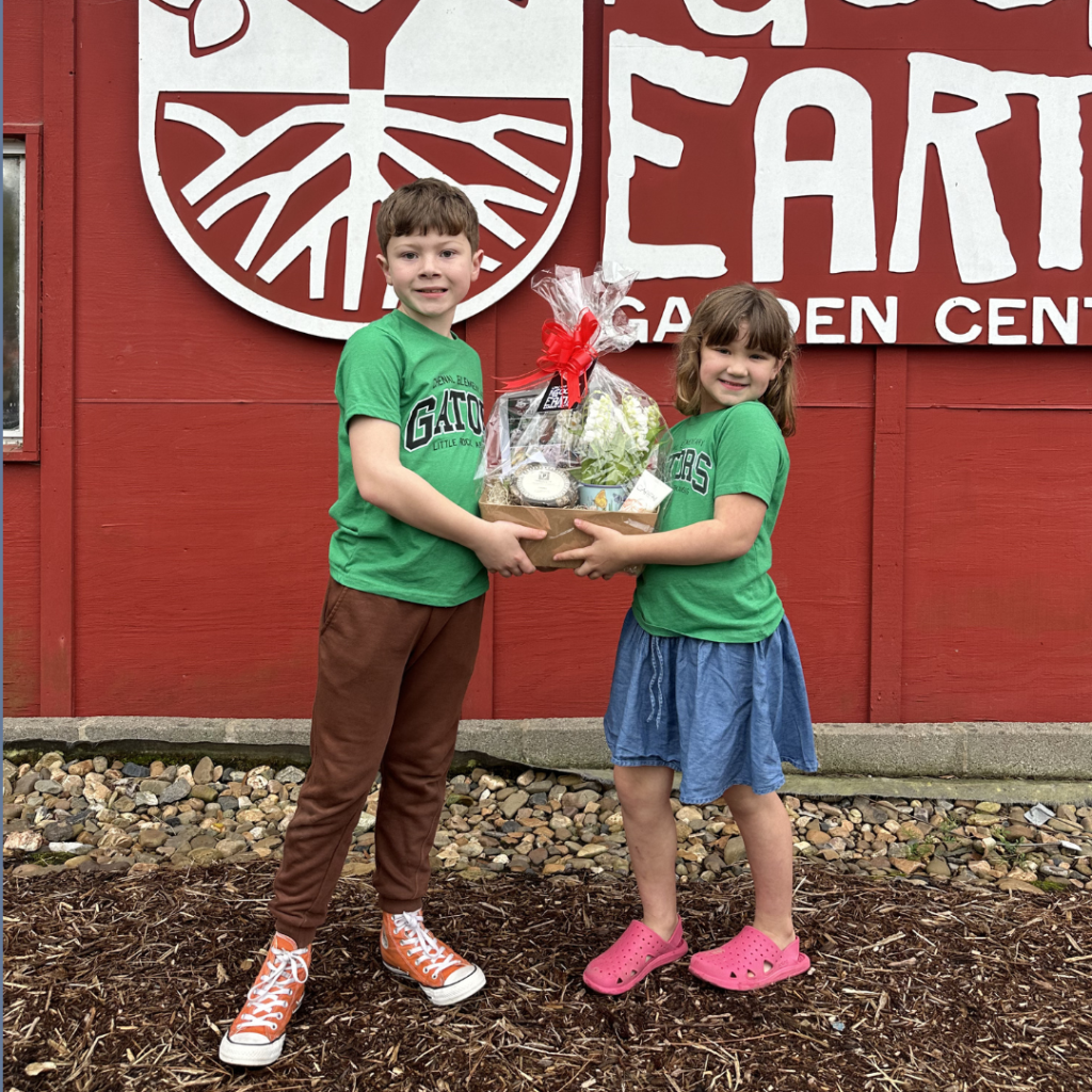 Two smiling children in green shirts and colorful shoes hold a gift basket together in front of a red building with a partial sign that reads EARTH. They stand on wood chips and gravel.