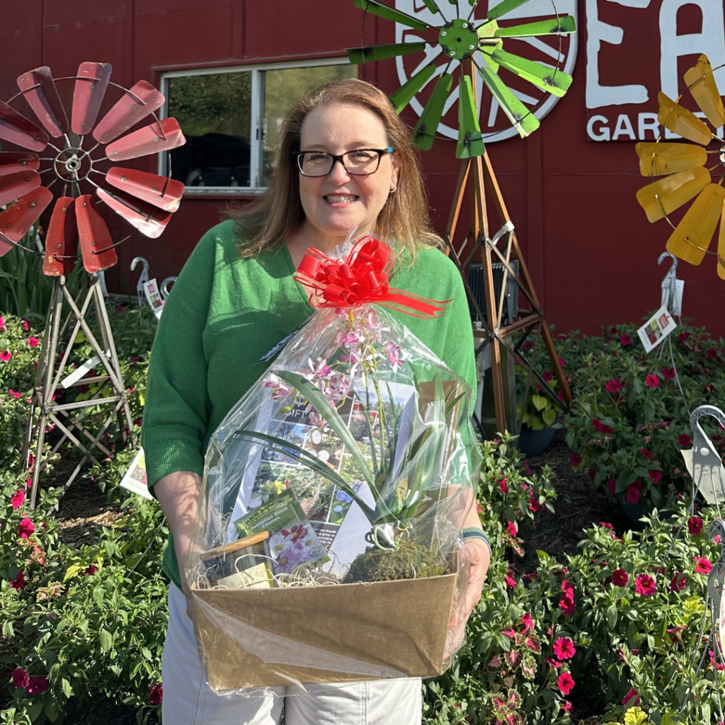 A woman in a green sweater holds a gift basket with a red bow, standing outside in front of colorful metal windmills and blooming flowers.