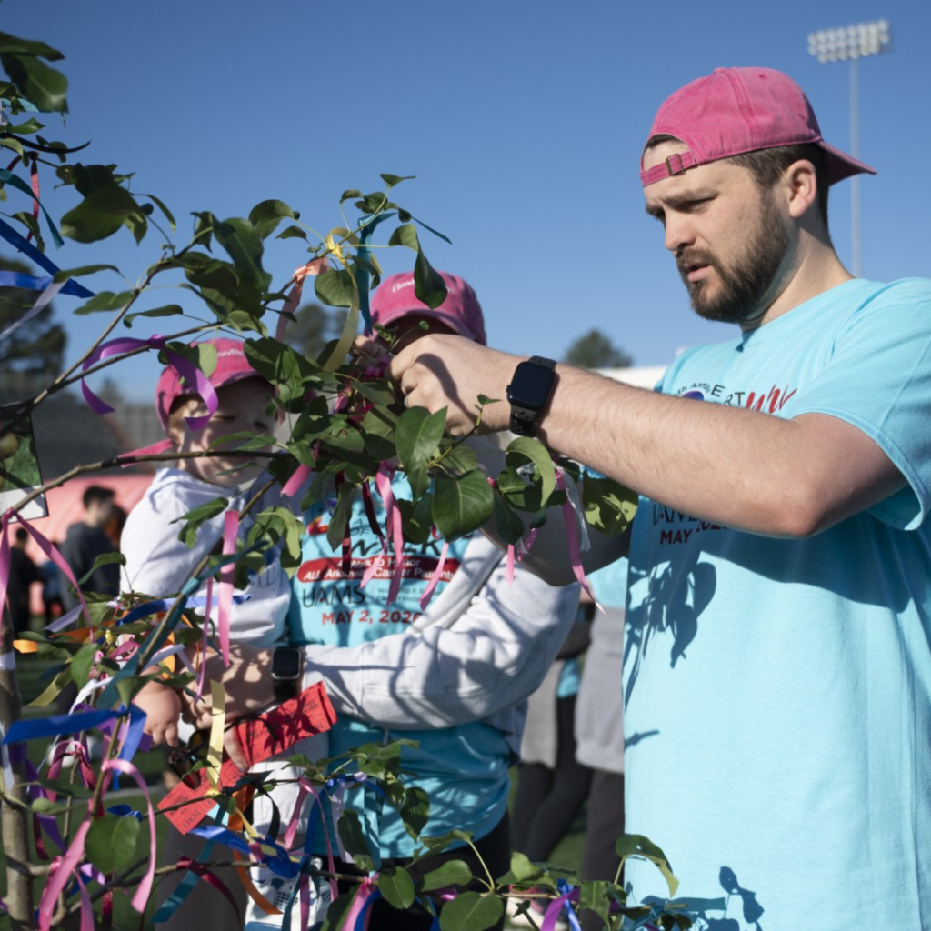 A man in a pink hat ties a ribbon to a tree branch at an outdoor event, while holding a child. Other people in matching shirts and hats are visible in the background under a clear blue sky.