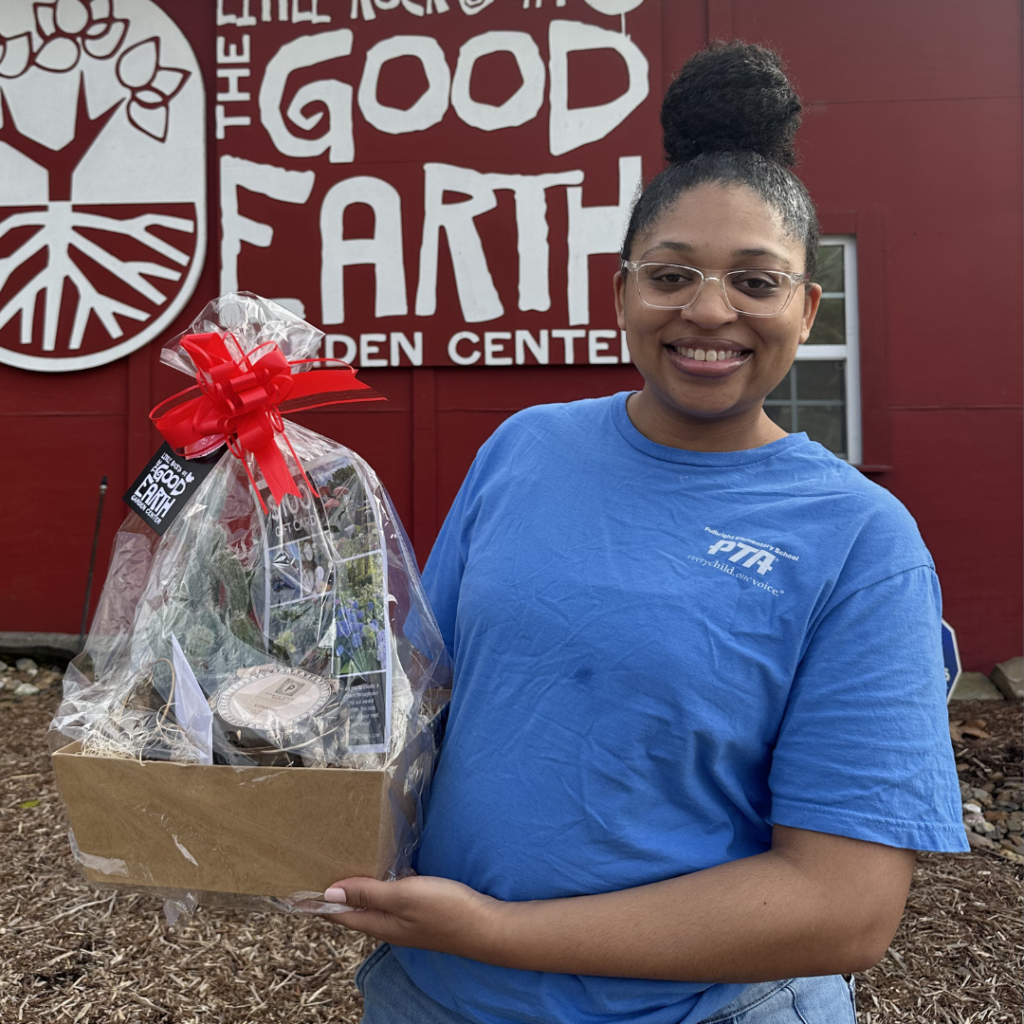 Woman in blue shirt holding a gift basket wrapped in plastic and red ribbon, smiling in front of a red building with a white “Good Earth” sign.