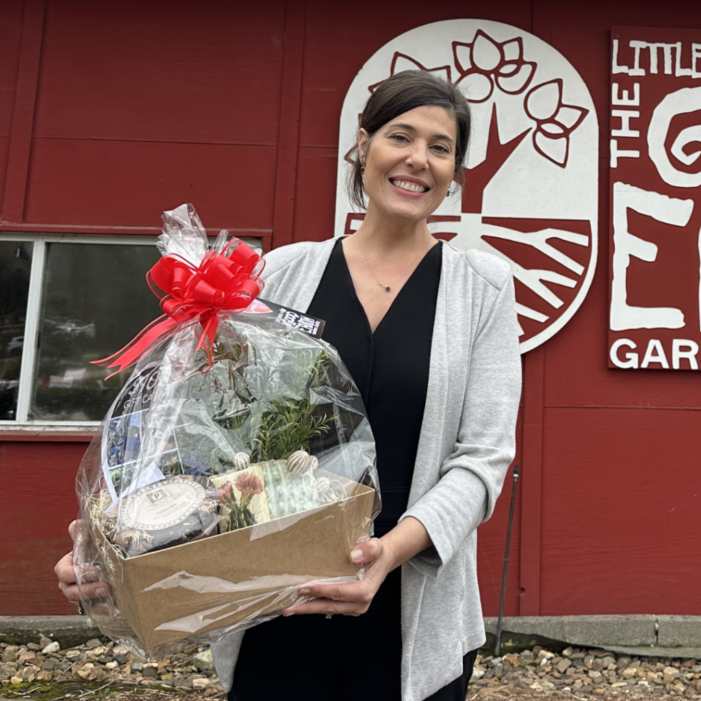 A smiling woman holds a gift basket with plants and treats, standing in front of a red building with a white floral logo and the words The Little Garden on the wall.