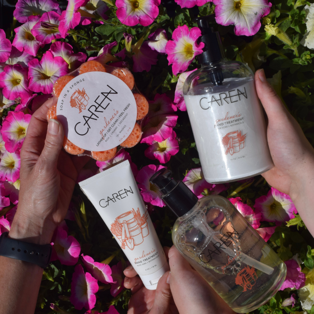 Four hands hold CareA skincare products—body wash, hand treatment, hand soap, and konjac sponge—over a background of vibrant pink and white flowers, creating a scene reminiscent of a beautifully curated gift shop display.