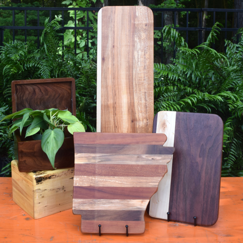 Five wooden boards and boxes of different shapes and colors, one holding a leafy green plant, are artfully arranged on an orange table outdoors with lush green ferns in the background—an inviting scene reminiscent of a stylish gift shop display.