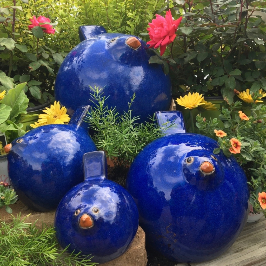 Four shiny blue ceramic bird sculptures are displayed among green plants and colorful flowers, including yellow daisies and pink roses, in a garden setting—creating a charming scene perfect for any gift shop display.