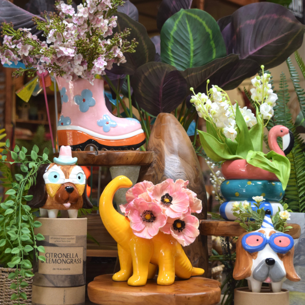 Colorful ceramic planters shaped like dogs, a yellow dinosaur with pink flowers, and a boot with blue flowers are surrounded by lush green and white plants, flowers, and citronella lemongrass candles in a decorative display perfect for any gift shop.