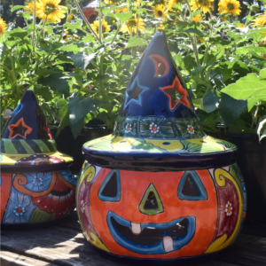 A colorful ceramic jack-o-lantern with a painted witch hat sits on a wooden table surrounded by yellow flowering plants in sunlight, creating the perfect cheerful display for a gift shop. Another similar ceramic piece is partially visible to the left.