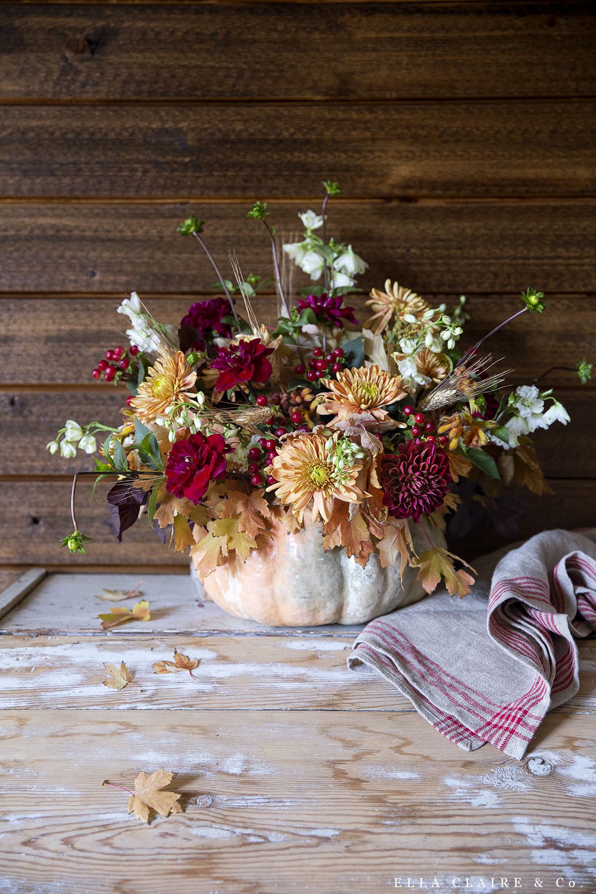 An autumn floral arrangement featuring dahlias and dried leaves displayed in a hollowed-out pumpkin on a weathered wooden table.