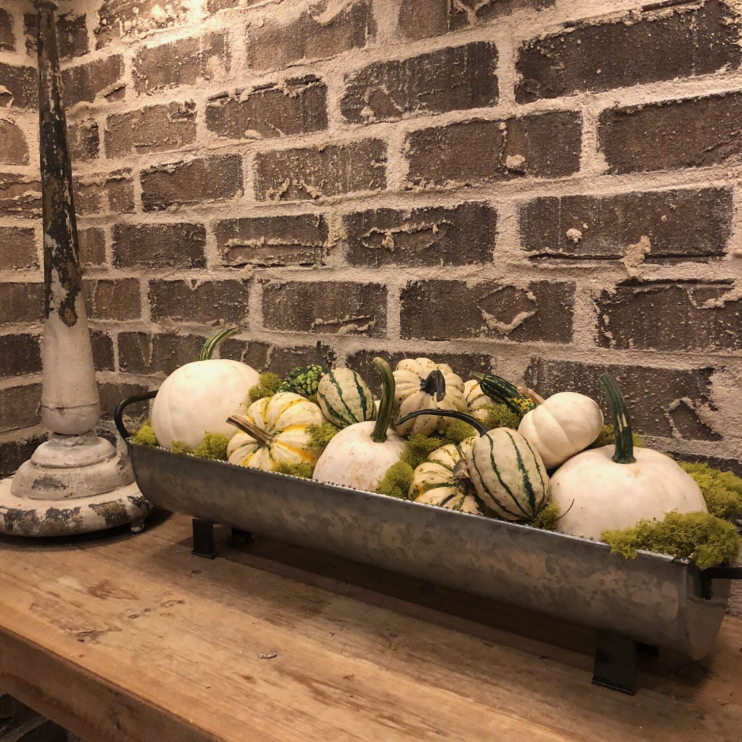 A rectangular metal trough filled with moss and various decorative gourds, placed on a wooden table in front of a brick wall.